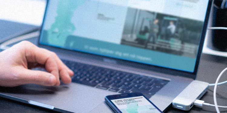 Närbild på hand vid dator och mobil som visar samma webbsida